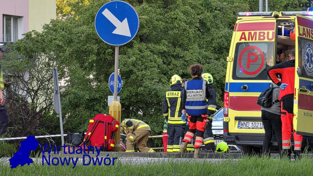 Grupa ratowników medycznych i strażaków pracuje przy wypadku drogowym, obok karetki i oznaczeń drogowych.