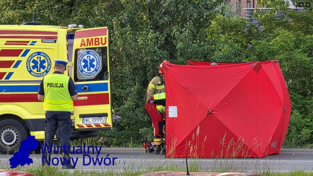 Ratownik medyczny w żółtym kombinezonie ustawia czerwony namiot przy ambulansie, a policjant w kamizelce obserwuje sytuację.