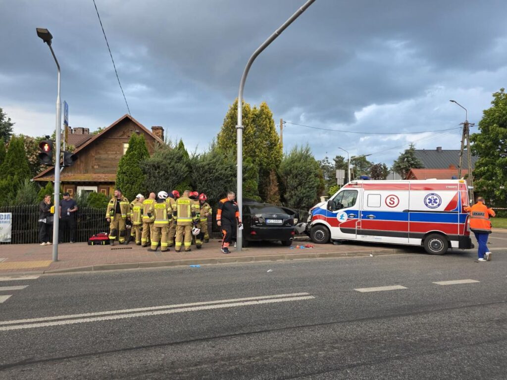 Grupa strażaków w mundurach omawia sytuację obok uszkodzonego samochodu, obok stoi ambulans.
