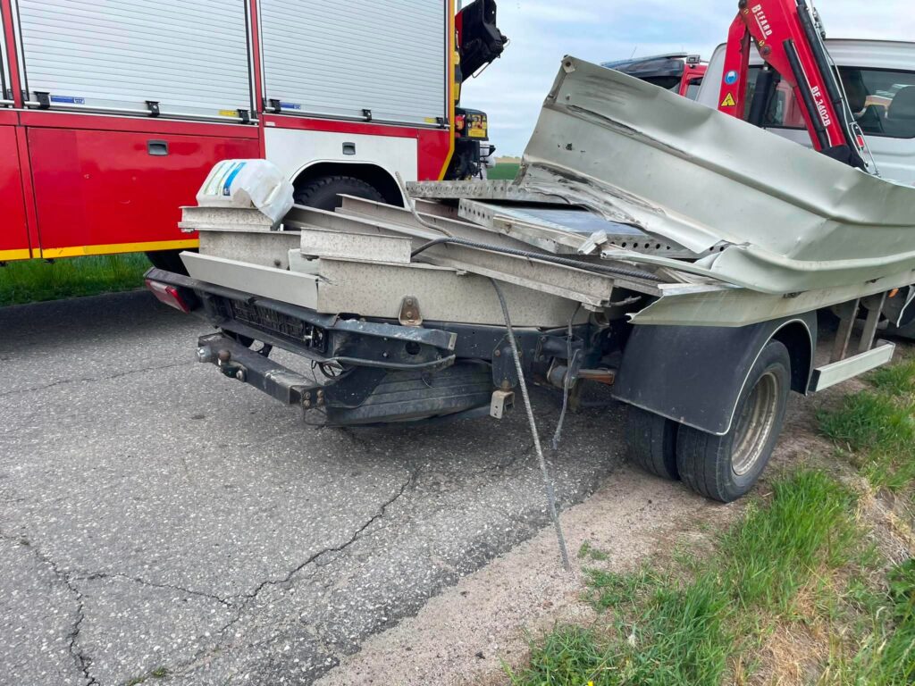 Uszkodzony samochód dostawczy z wygiętą skrzynią ładunkową stoi na poboczu drogi, obok wozu strażackiego.