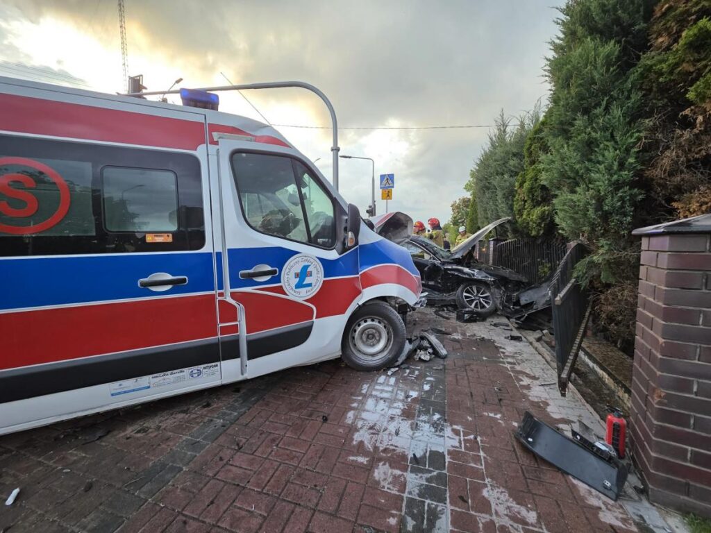 Karetka pogotowia stojąca na chodniku obok zderzonego samochodu osobowego, w tle widoczni strażacy.