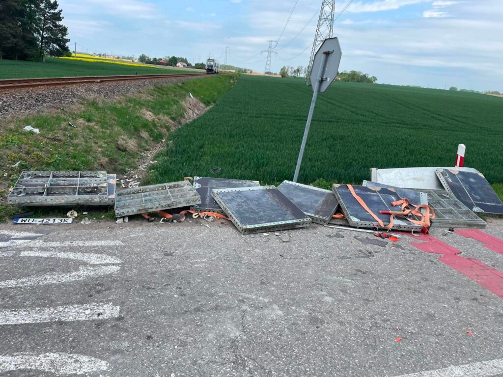 Zestaw metalowych paneli leżących na drodze obok torów kolejowych, w tle zielone pole.