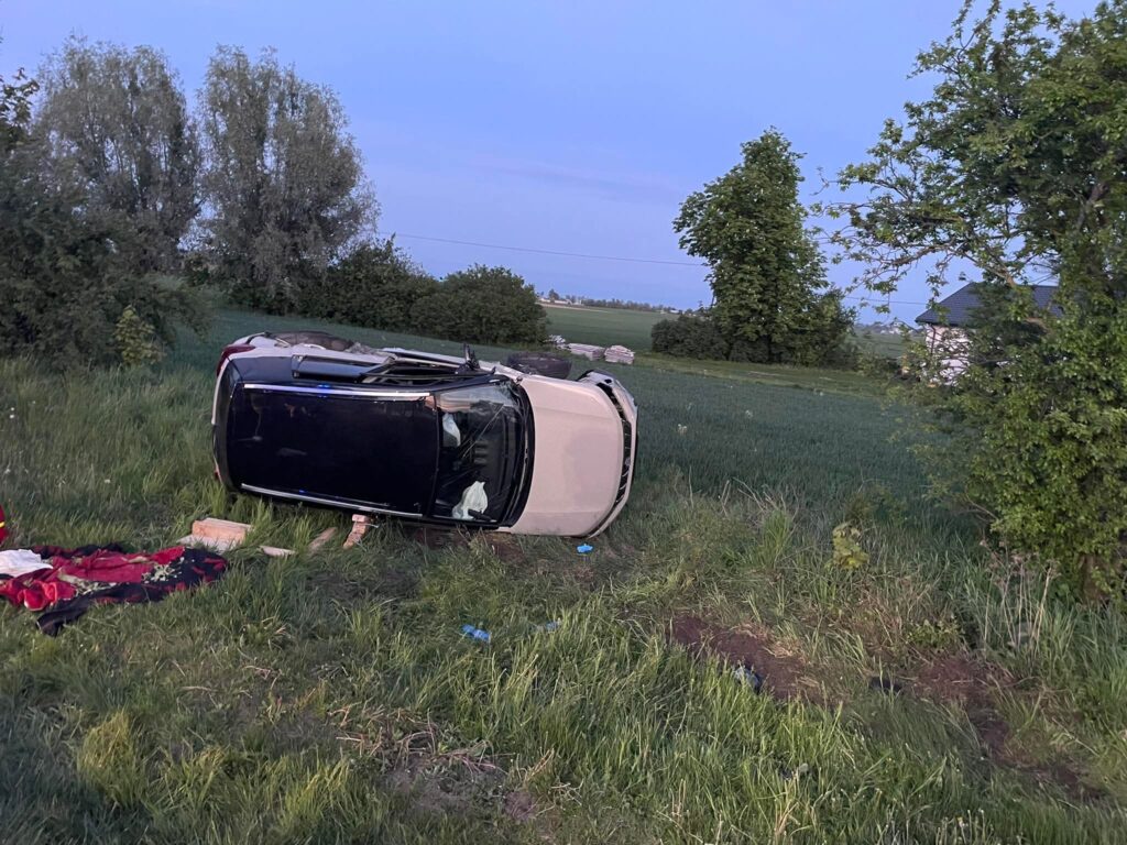 Przewrócone auto leży na poboczu drogi wśród zielonych pól, obok widoczna koc i kilka przedmiotów.