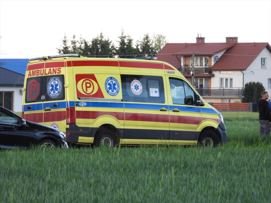 Karetka pogotowia zaparkowana na polu, obok stoi dwóch mężczyzn rozmawiających ze sobą.