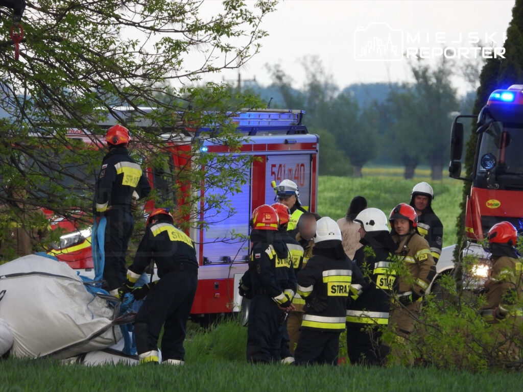Grupa strażaków w mundurach pracuje przy wraku balonu, obok stoją dwa samochody strażackie w zielonym otoczeniu.