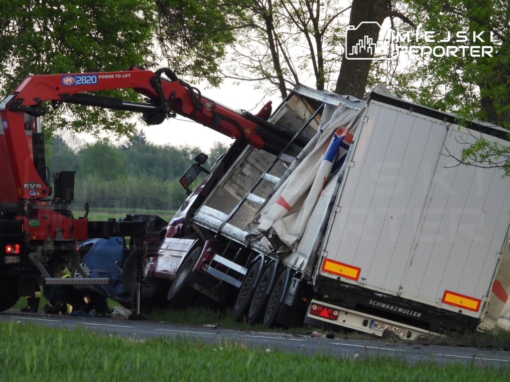 Czerwony dźwig podnosi przewróconą ciężarówkę na poboczu drogi, otoczoną zielenią i drzewami.