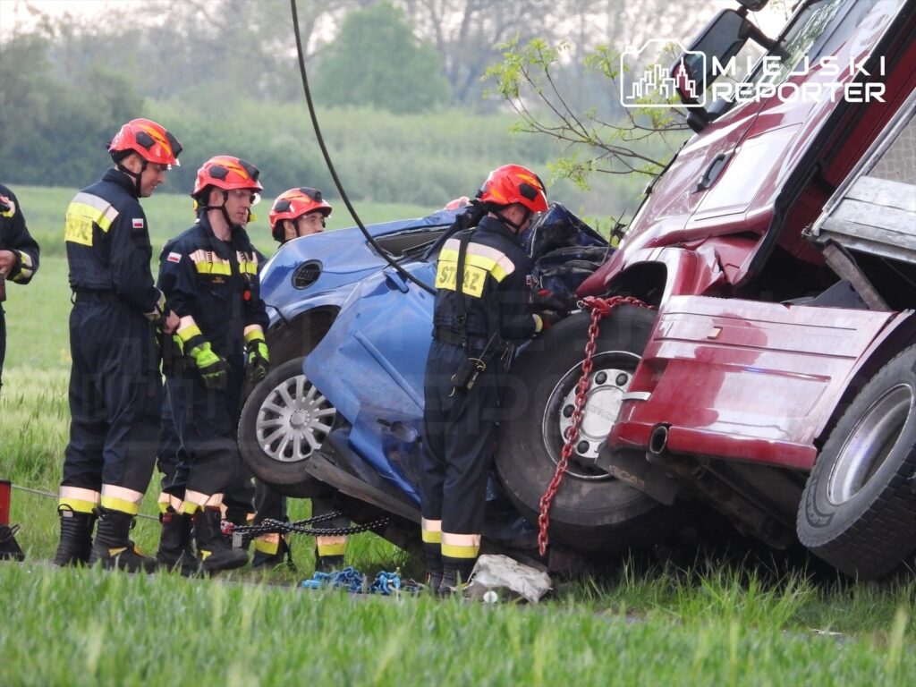 Grupa strażaków w kaskach pracuje nad wydobyciem niebieskiego samochodu osobowego z pod ciężarówki na polu.