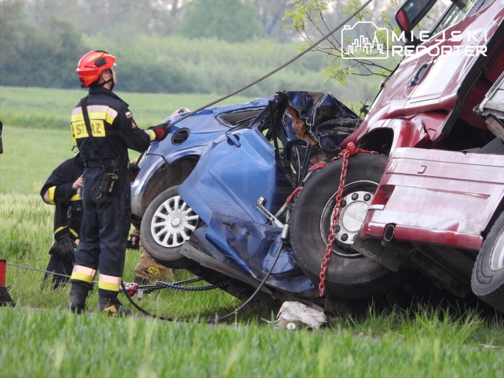 Strażacy pracują nad wydobyciem poszkodowanego z niebieskiego samochodu osobowego, który zderzył się z ciężarówką.