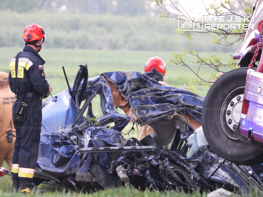 Strażak w hełmie bada wrak rozbitego samochodu na tle zielonego krajobrazu.