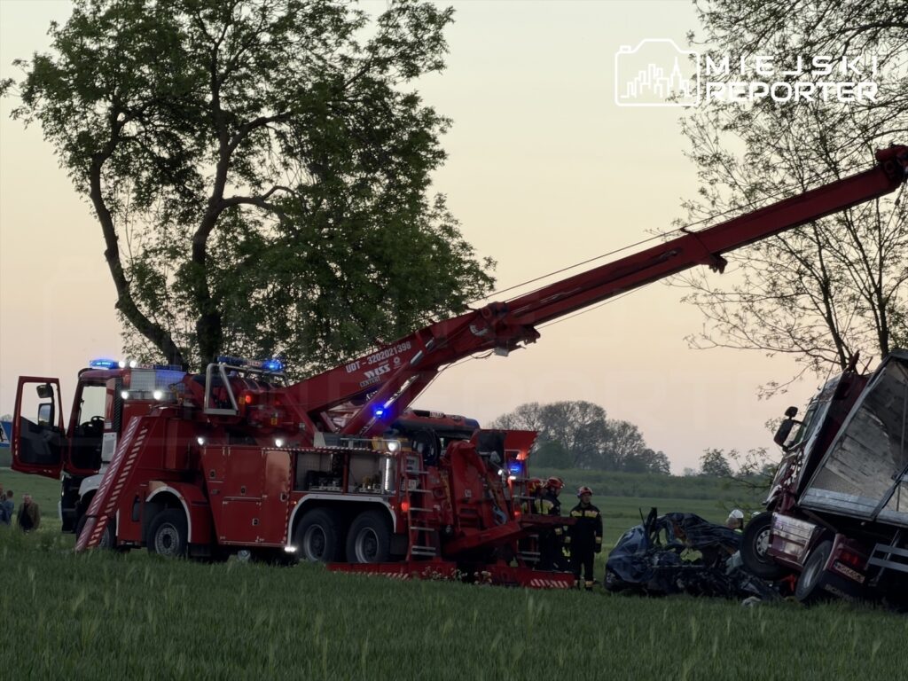 Czerwony wóz strażacki z podnośnikiem podnosi wrak ciężarówki w polu, obok pracują strażacy.