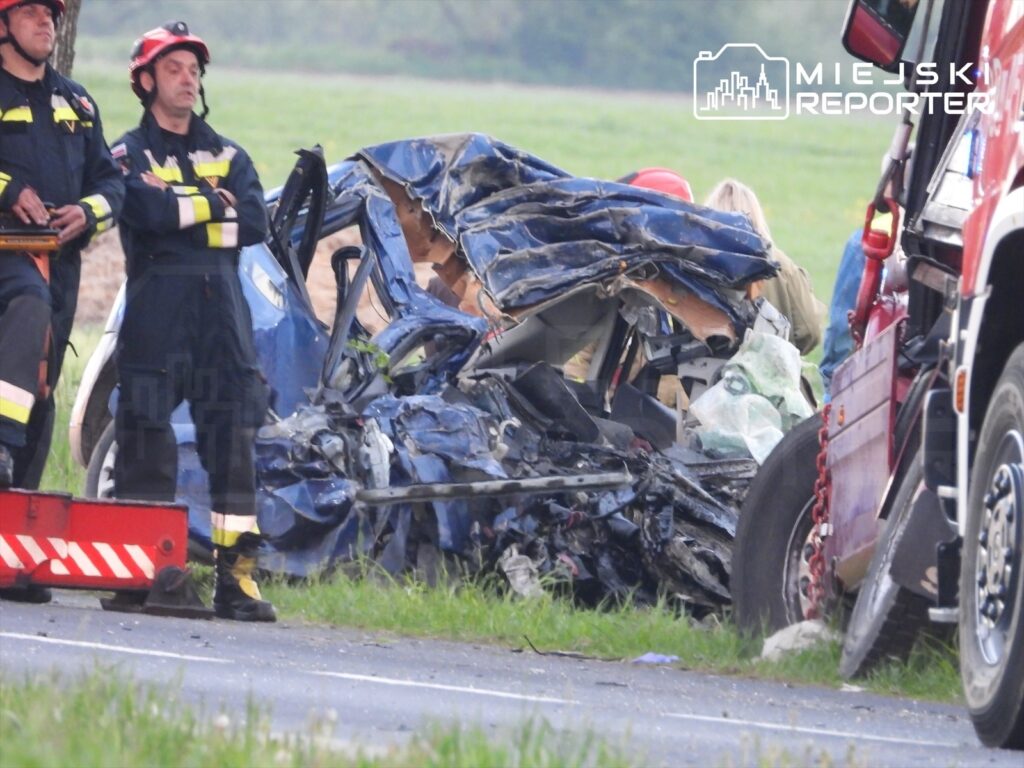 Dwaj strażacy w mundurach obserwują wrak niebieskiego samochodu osobowego po wypadku drogowym na poboczu.