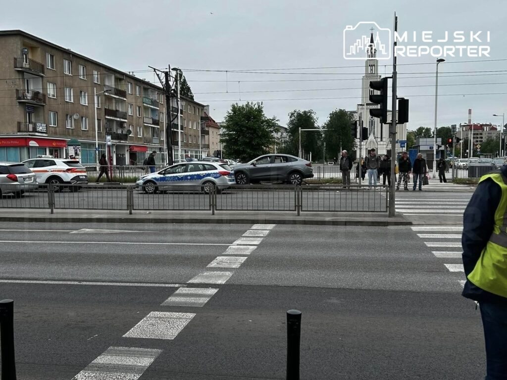 Grupa osób czeka na przejściu dla pieszych w pobliżu skrzyżowania, obok radiowozów policyjnych i budynków mieszkalnych.