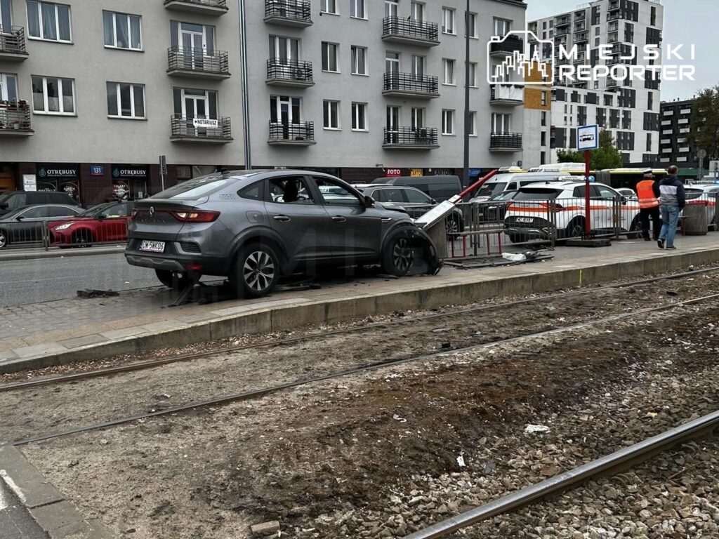 Zniszczony samochód osobowy stojący na chodniku obok torów tramwajowych, przy nowoczesnym budynku mieszkalnym.