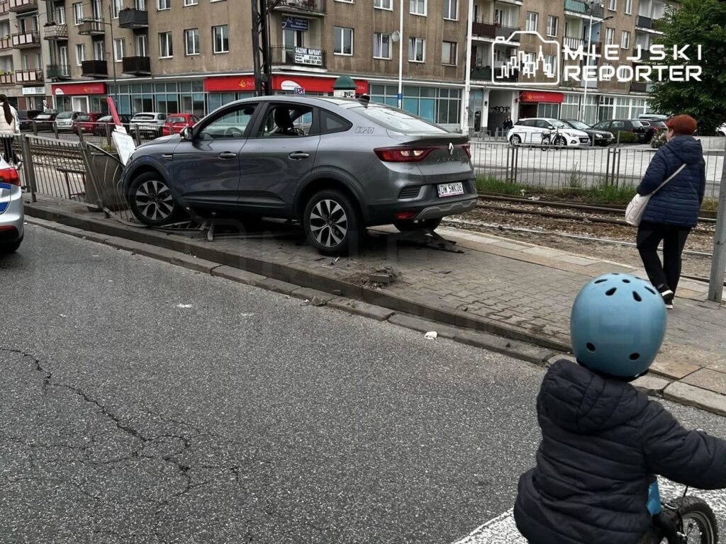 Szary samochód osobowy zaparkowany na chodniku obok torów tramwajowych, obok przechodząca kobieta i dziecko na rowerze.