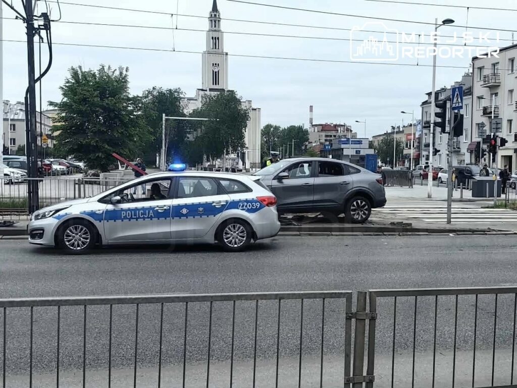 Policyjny radiowóz z włączonymi sygnałami świetlnymi stoi obok uszkodzonego samochodu na skrzyżowaniu w mieście.