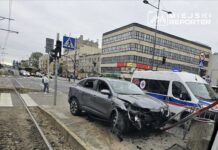 Uszkodzony samochód osobowy stoi obok przystanku tramwajowego, a obok niego znajduje się karetka pogotowia.
