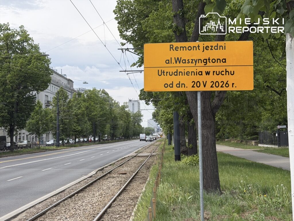 Znak drogowy informujący o remoncie jezdni al. Waszyngtona oraz utrudnieniach w ruchu od 20 maja 2026 roku, w otoczeniu drzew i budynków.