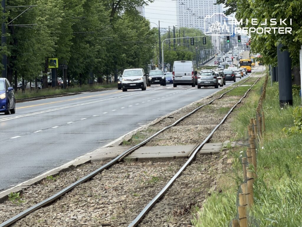 Ruchliwa ulica z samochodami, obok torów tramwajowych i zielonymi drzewami wzdłuż jezdni.