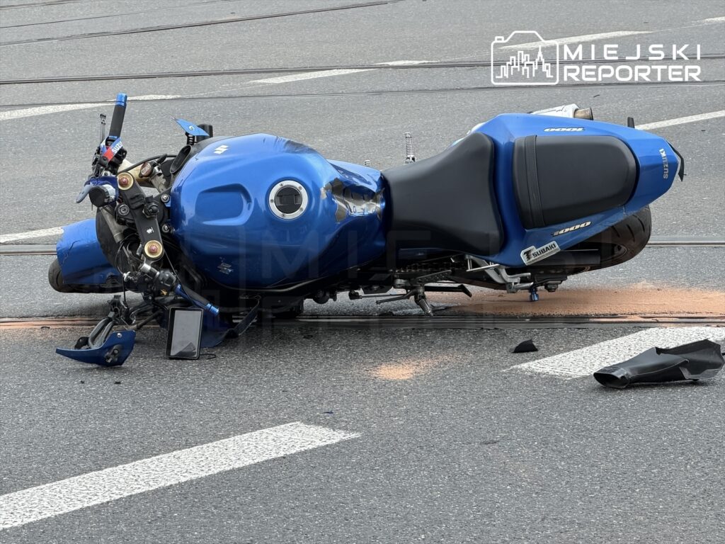 Nieprzewrócony niebieski motocykl leży na jezdni, obok widoczne fragmenty uszkodzonego plastiku.