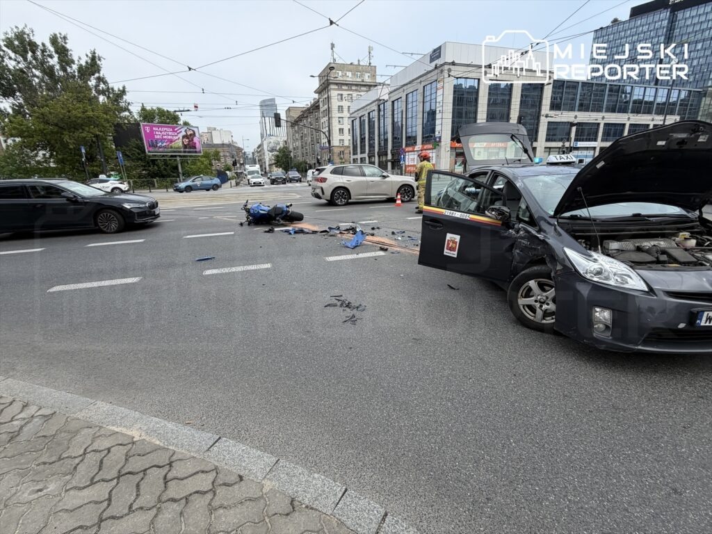 Motocykl leży na jezdni obok uszkodzonego samochodu osobowego w miejskim otoczeniu, wokół ruch uliczny.