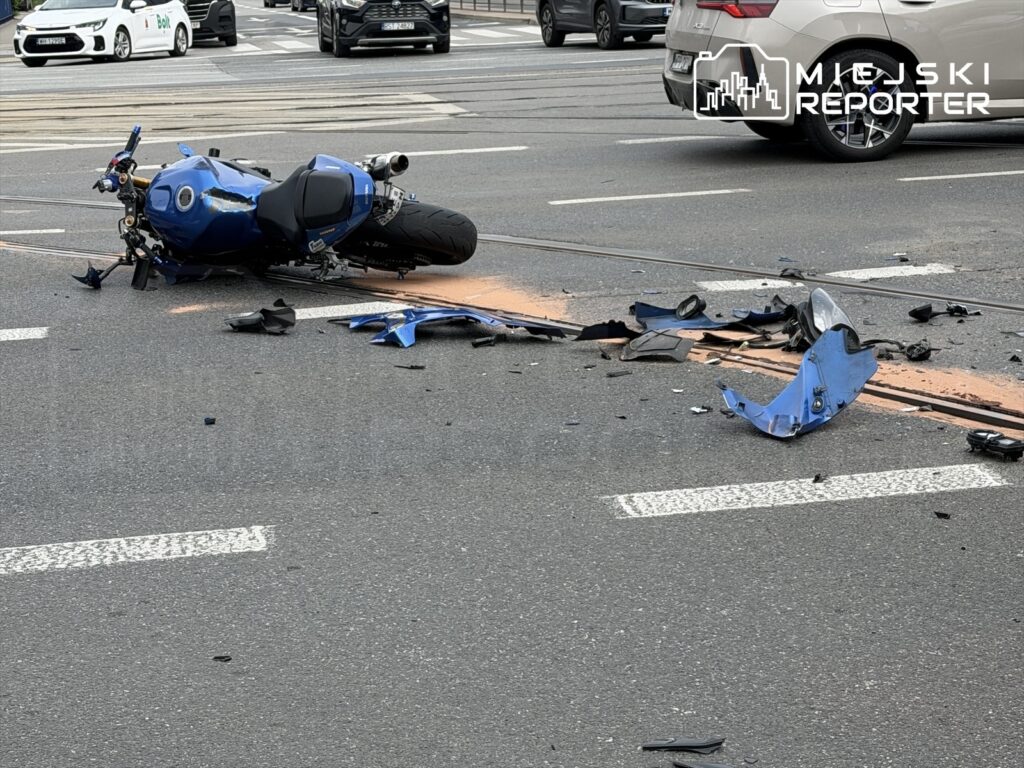 Motocykl leży na jezdni, otoczony rozbitymi elementami, w ruchliwej miejskiej scenerii.