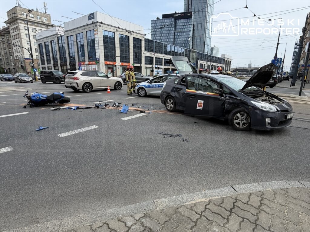 Motocykl leży na jezdni obok uszkodzonego samochodu osobowego z otwartą maską, a w tle widać budynki biurowe.