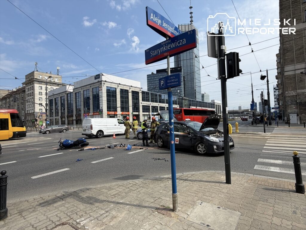 Na ulicy, w pobliżu przystanku tramwajowego, ratownicy udzielają pomocy poszkodowanym po wypadku drogowym. Leży motor, a samochód ma otwartą maskę. W tle nowoczesne budynki i tramwaj.