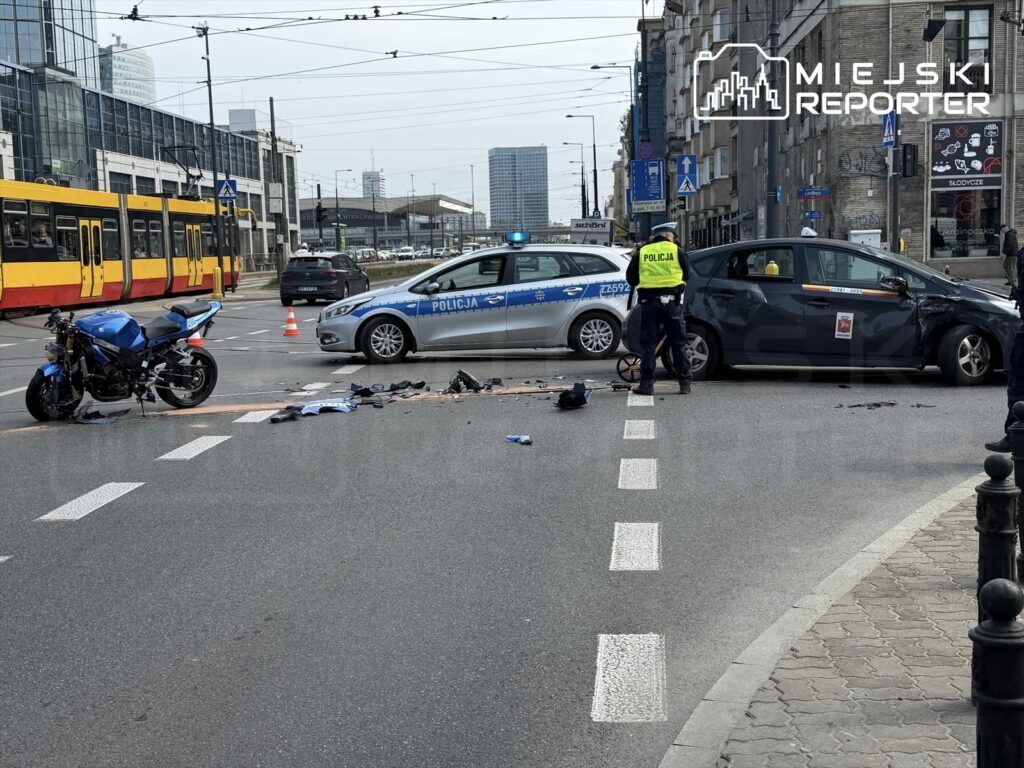 Motocykl leży na drodze obok policyjnego radiowozu, a funkcjonariusz zabezpiecza miejsce wypadku. W tle widać tramwaj.