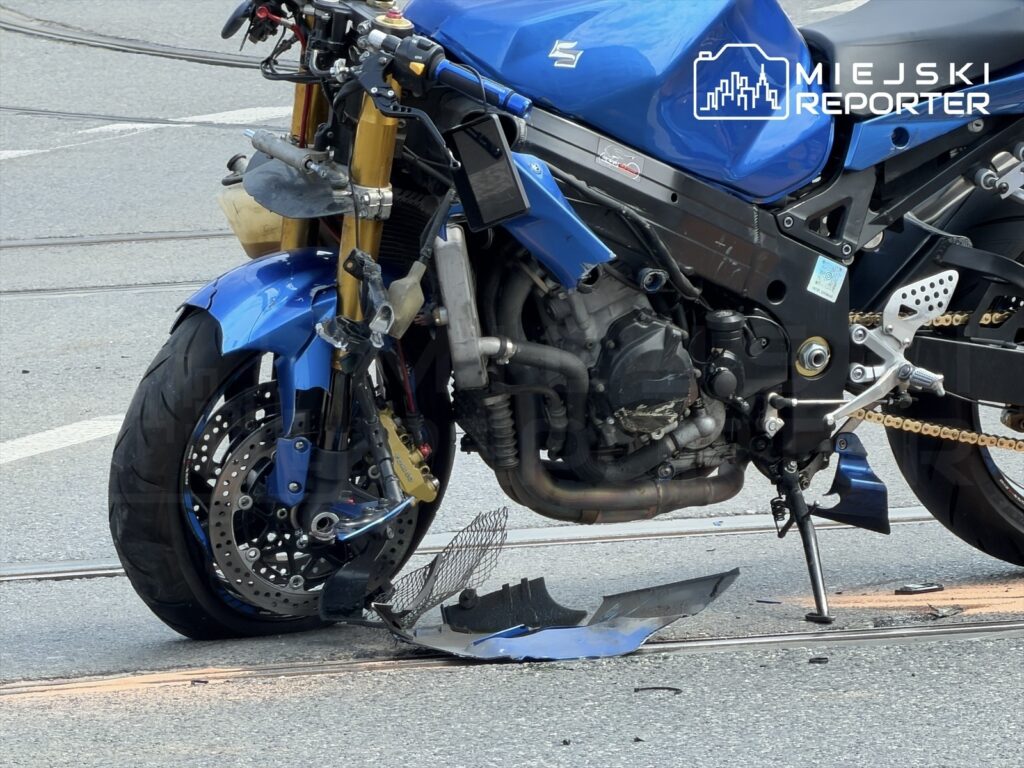 Uszkodzony niebieski motocykl stoi na torach tramwajowych, obok leżące fragmenty karoserii.
