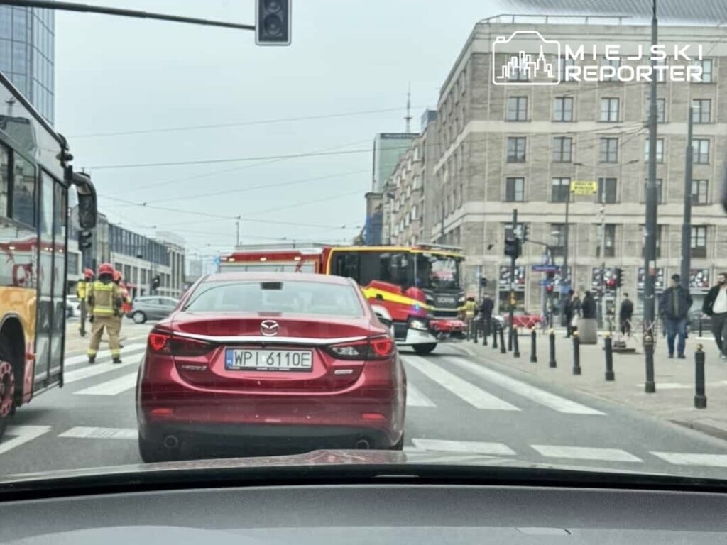 Czerwony samochód osobowy zatrzymany na pasach, obok strażacy i pojazd strażacki w miejskim otoczeniu.