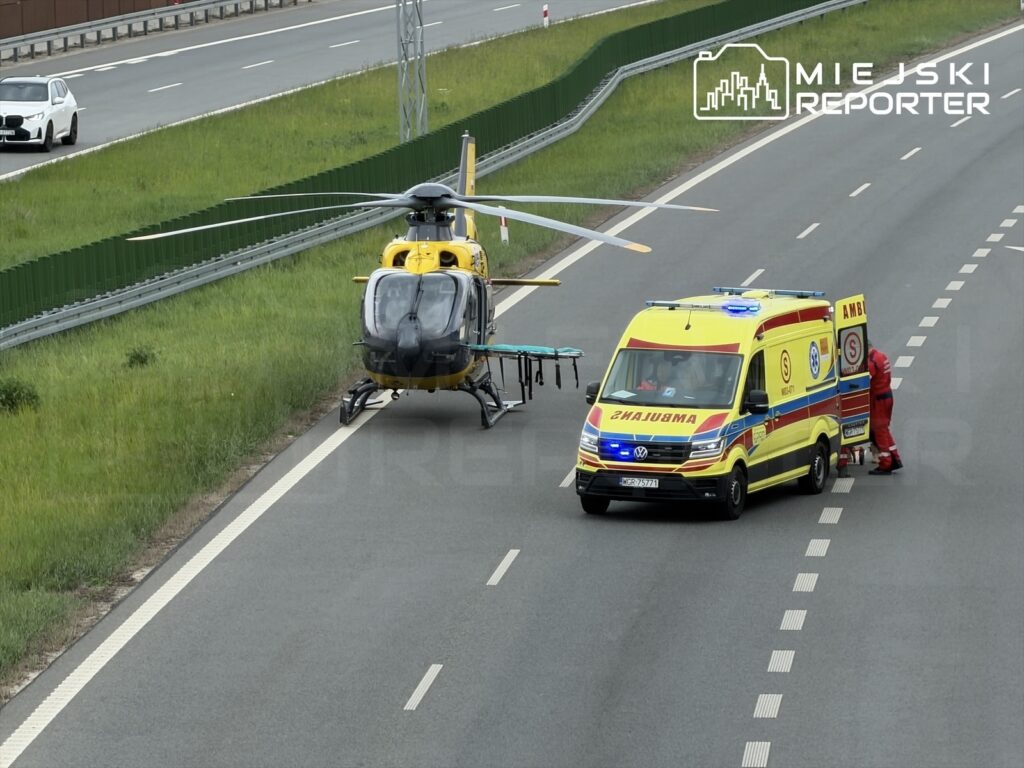 Helikopter ratunkowy ląduje obok karetki na autostradzie, gdzie ratownicy przygotowują nosze do transportu.