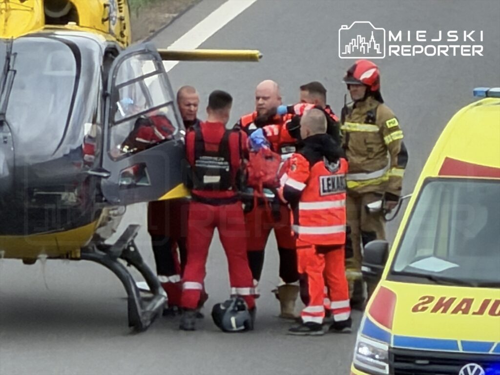 Grupa ratowników w czerwonych strojach omawia działania przy helikopterze ratunkowym na drodze.