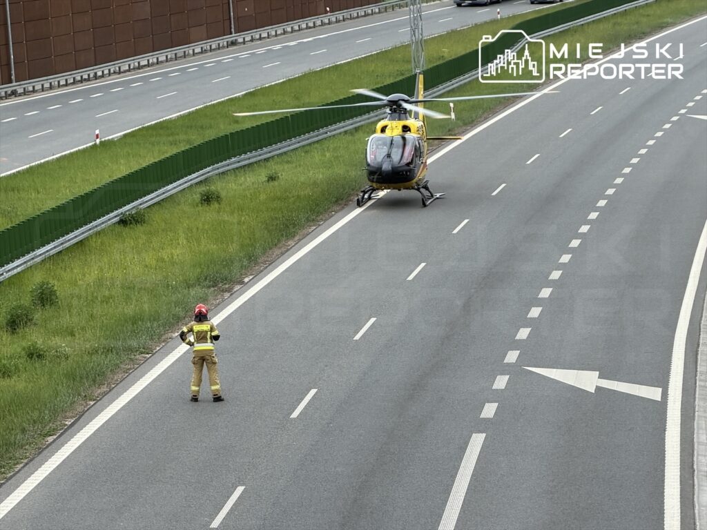 Strażak w hełmie kieruje ruchem obok lądowiska dla helikoptera na poboczu drogi.