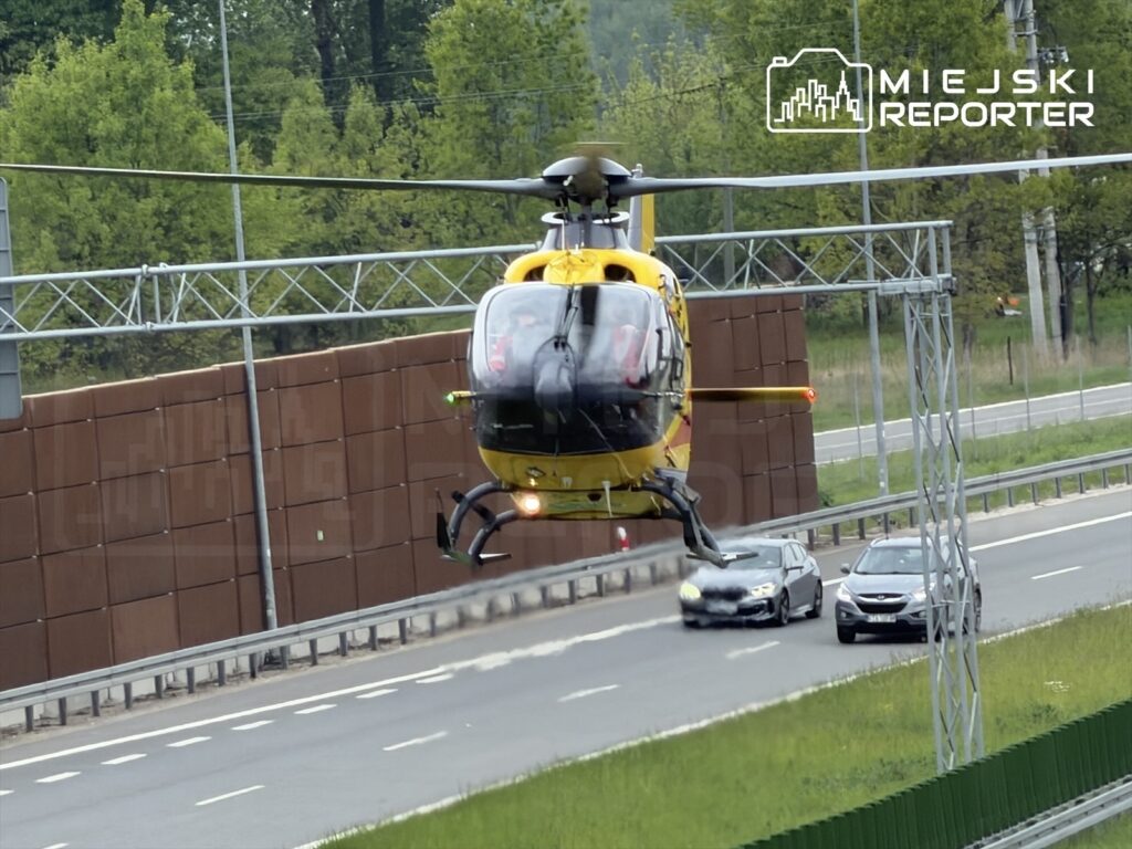 Helikopter ratunkowy unosi się nad ruchem samochodowym na drodze, w tle widoczne drzewa i ogrodzenie.