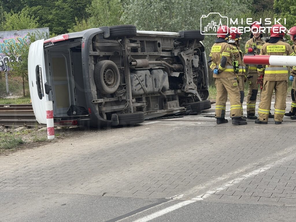 Przewrócony furgon leży na boku obok torów kolejowych, a strażacy w mundurach omawiają sytuację.
