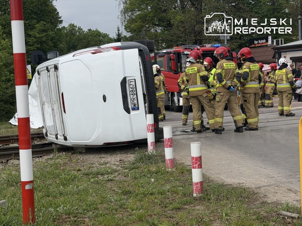 Grupa strażaków w mundurach, pracująca przy przewróconym białym pojeździe obok torów kolejowych.