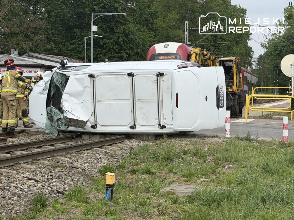 Biały samochód dostawczy leży na boku obok torów kolejowych, a strażacy pracują nad usunięciem pojazdu.