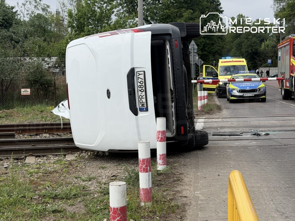 Przewrócony biały pojazd dostawczy leży na boku obok torów kolejowych, w tle widoczne radiowozy i karetki.