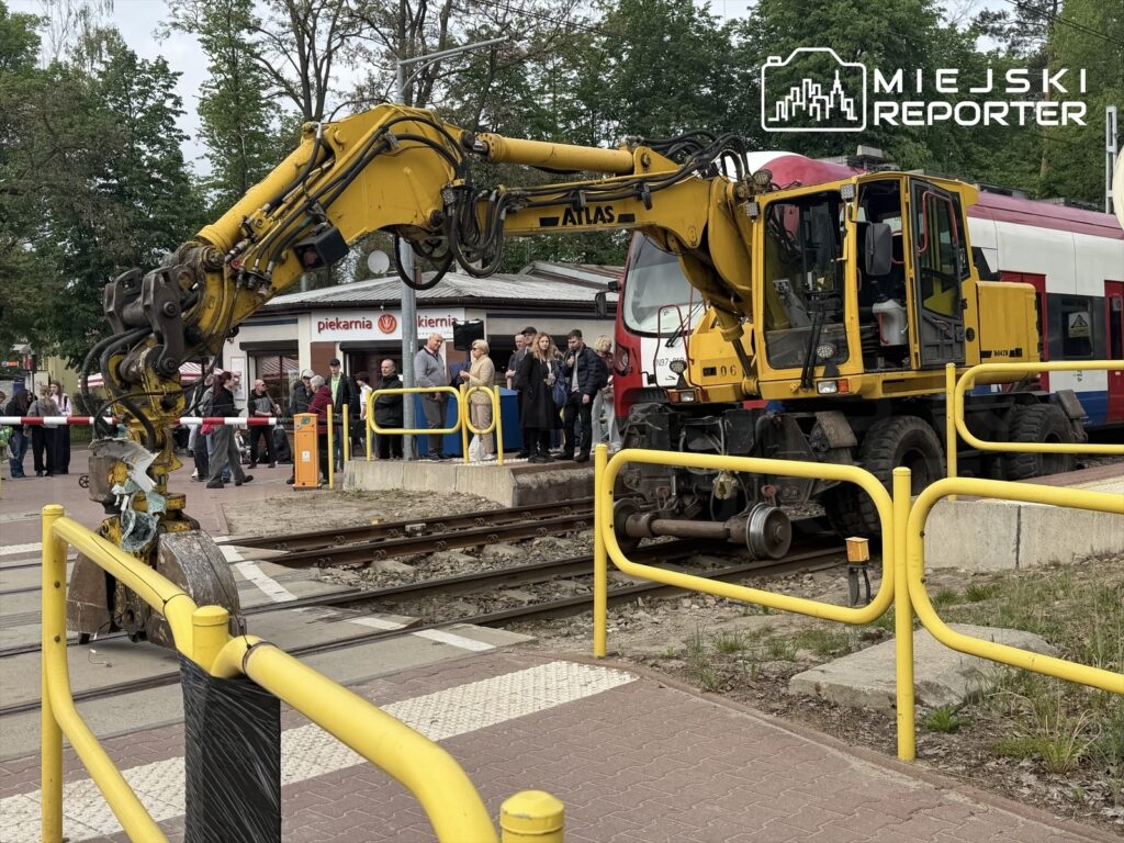 Maszyna budowlana z żółtym ramieniem pracuje na torach kolejowych, obok grupy ludzi obserwujących z zainteresowaniem.