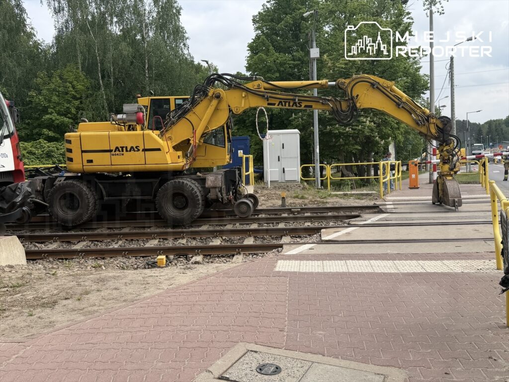 Żółty pojazd budowlany z wysięgnikiem pracuje na torach kolejowych obok chodnika, otoczony zielenią.