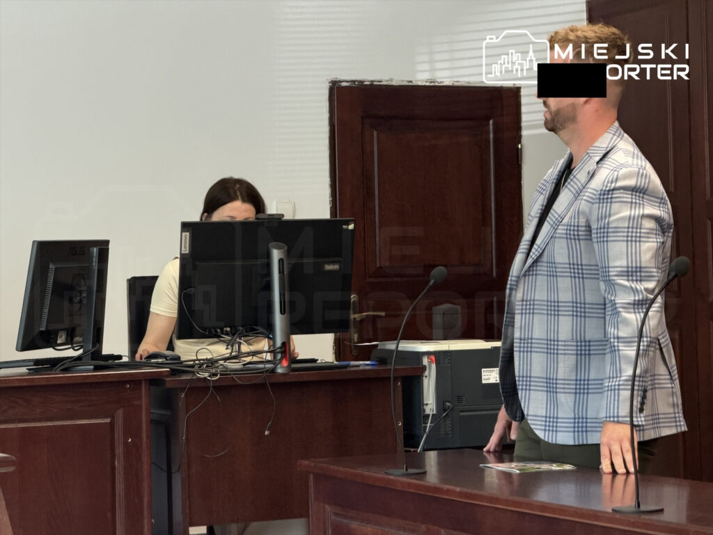 Mężczyzna w eleganckim garniturze stoi przy mównicy, podczas gdy kobieta pracuje przy dwóch komputerach w tle.