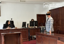 Mężczyzna w eleganckim garniturze stoi przed ławą sędziowską w sali sądowej, podczas rozprawy.