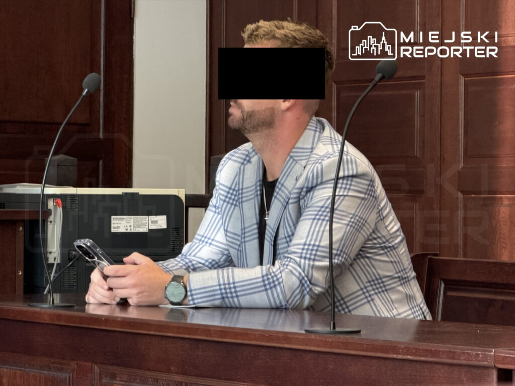 Mężczyzna w kratkowanym garniturze siedzi przy stole w sądzie, trzymając telefon w dłoniach.
