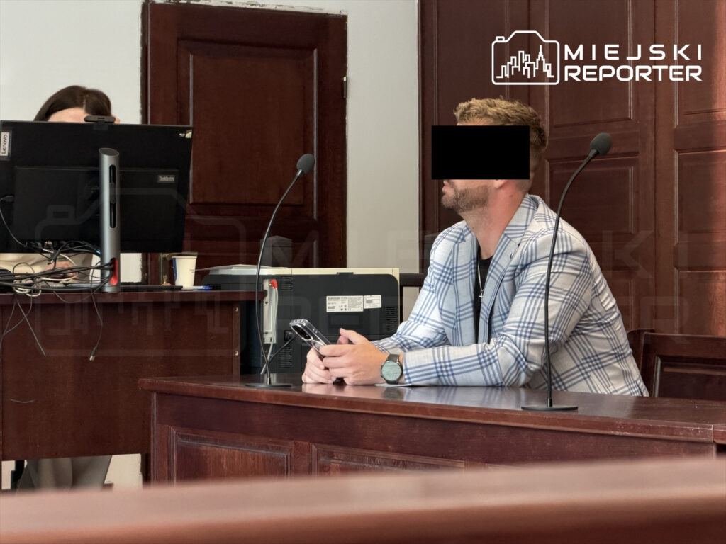Mężczyzna w jasnym garniturze siedzi przy stole w sali sądowej, trzymając telefon komórkowy.