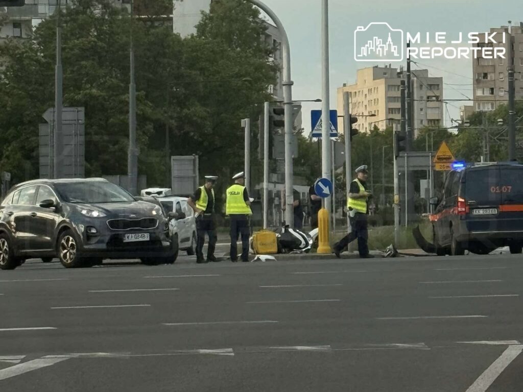 Policjanci w kamizelkach odblaskowych prowadzą czynności na skrzyżowaniu, obok uszkodzonego motocykla.