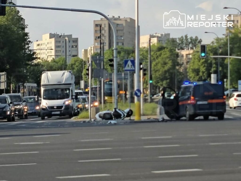 Motocykl leży na jezdni obok słupa, a w tle widać ruchliwe skrzyżowanie z zapalonymi światłami.