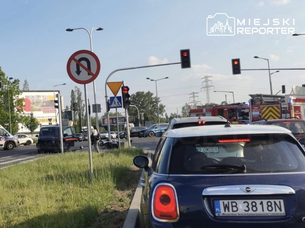 Kierowcy zatrzymani na czerwonym świetle przed skrzyżowaniem, w tle widoczny znak zakazu zawracania i radiowozy.