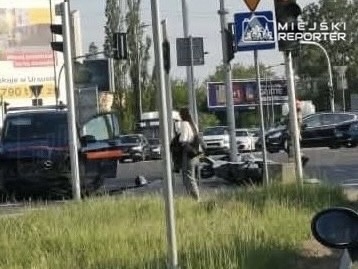 Motocyklista leży na jezdni obok przewróconego motocykla, a obok zatrzymują się samochody na ruchliwej ulicy.
