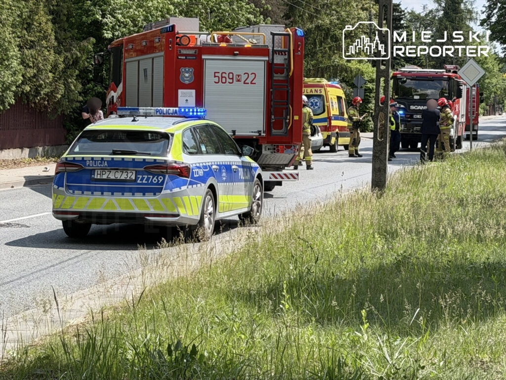 Na drodze stoi radiowóz policyjny z włączonymi sygnałami świetlnymi, obok niego zespół strażaków i ambulans.