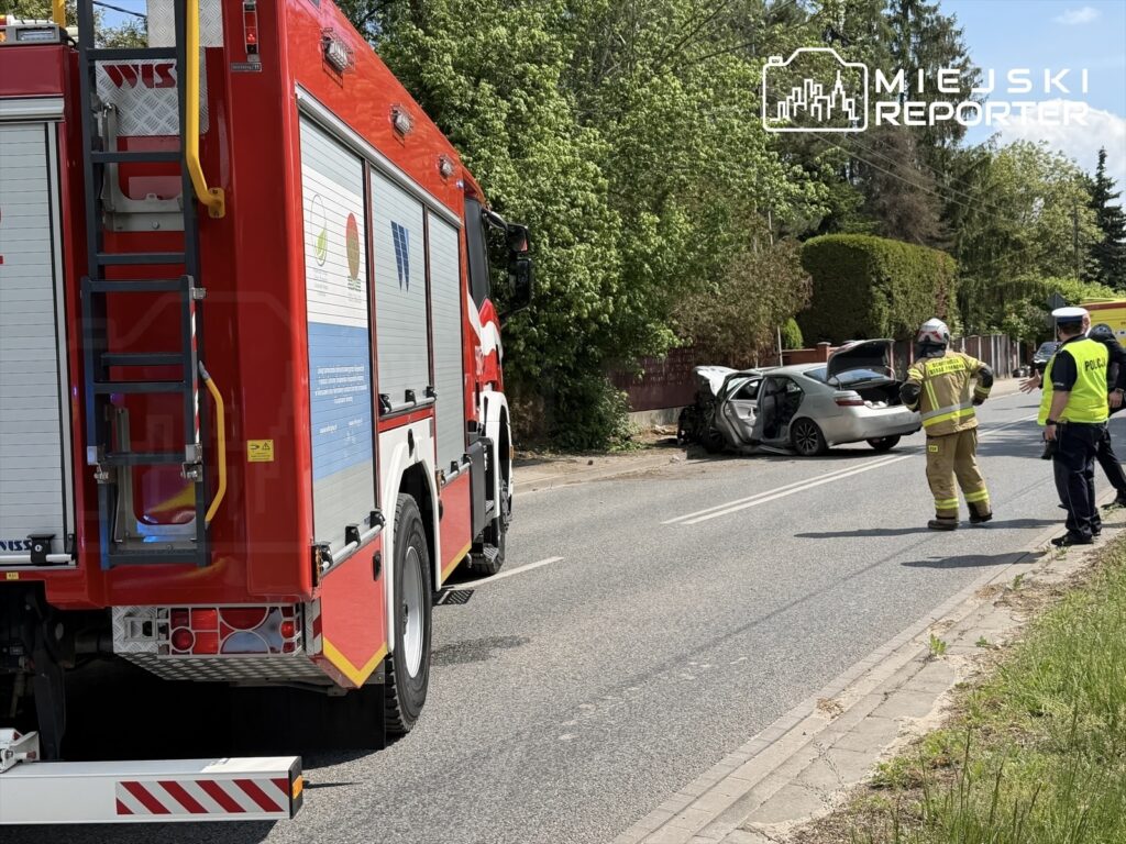 Czerwony wóz strażacki stoi na drodze obok uszkodzonego samochodu, w pobliżu pracują strażacy i policjanci.